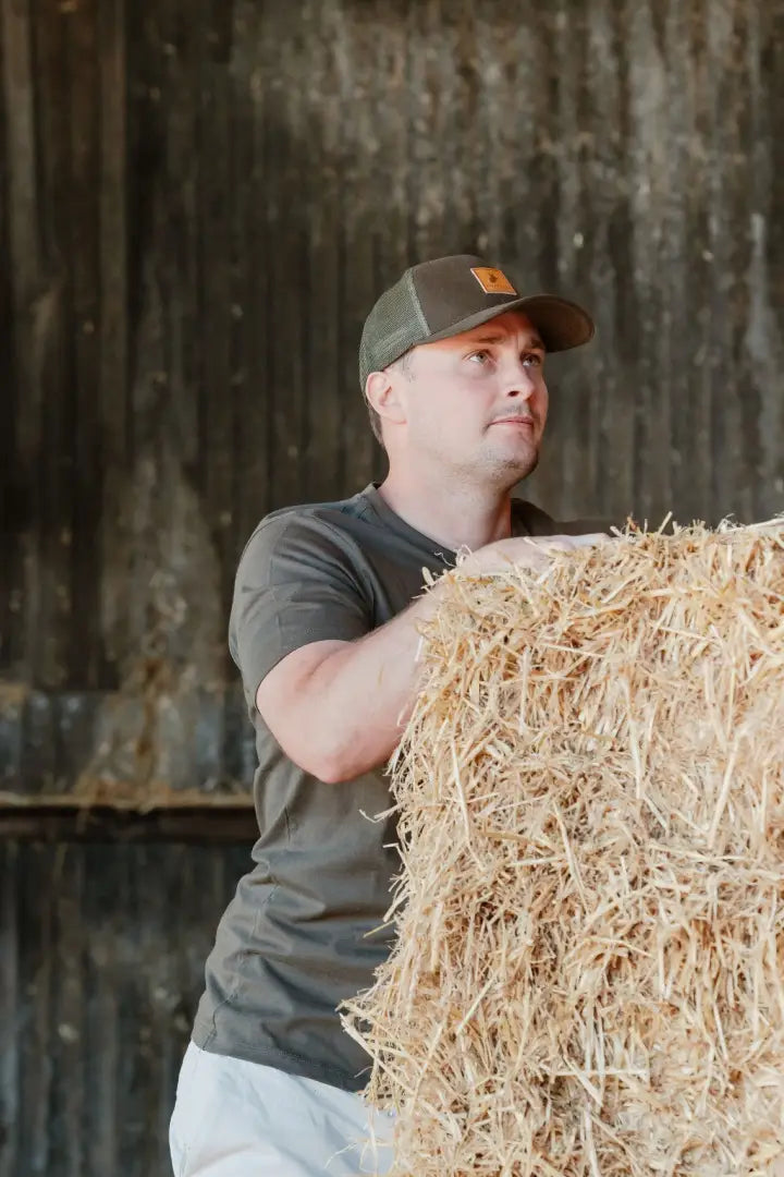 Man holding hay bale wearing New Forest Country Sport iconic trucker hat in outdoor farm scene
