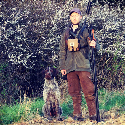 Hunter with a rifle and dog in flowering bushes wearing a New Forest Hawker Jacket